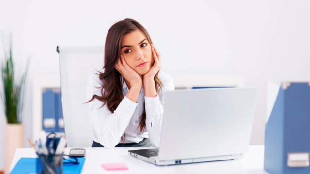 a girl with her head on her hands with a bored look on her face sitting at a work desk with a computer