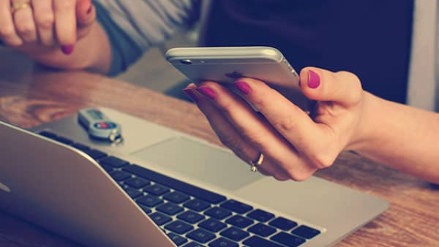 a closeup of a woman's hand holding an iPhone while sitting in front of a laptop